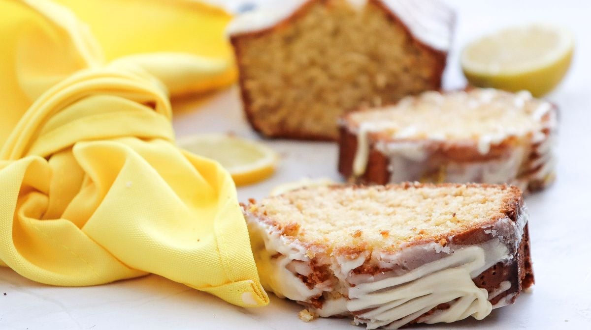 Two slices of glazed lemon loaf cake sit on a white surface with a yellow cloth, lemon slices, and the remaining loaf in the background.