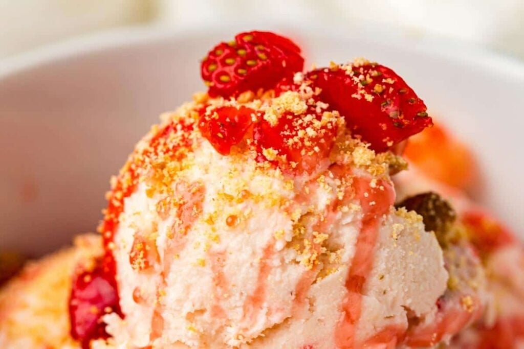 A close-up of a bowl of strawberry ice cream topped with strawberry pieces, crushed crumbs, and strawberry syrup.