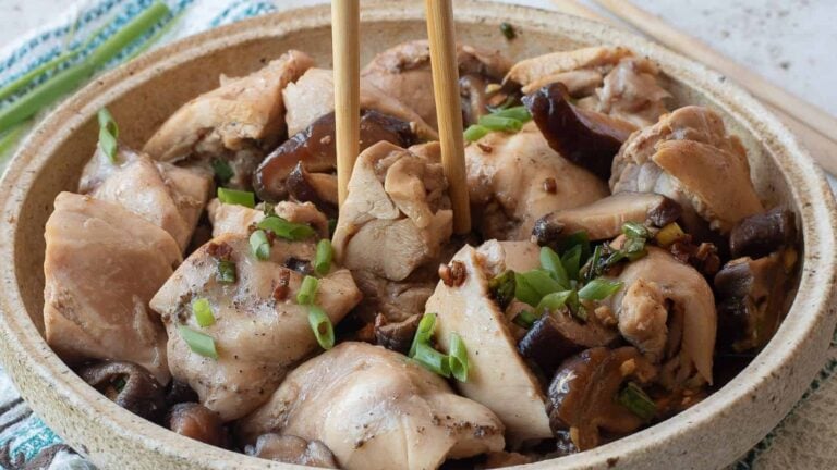 A bowl of steamed chicken pieces with mushrooms, garnished with chopped green onions, with chopsticks picking up a piece.