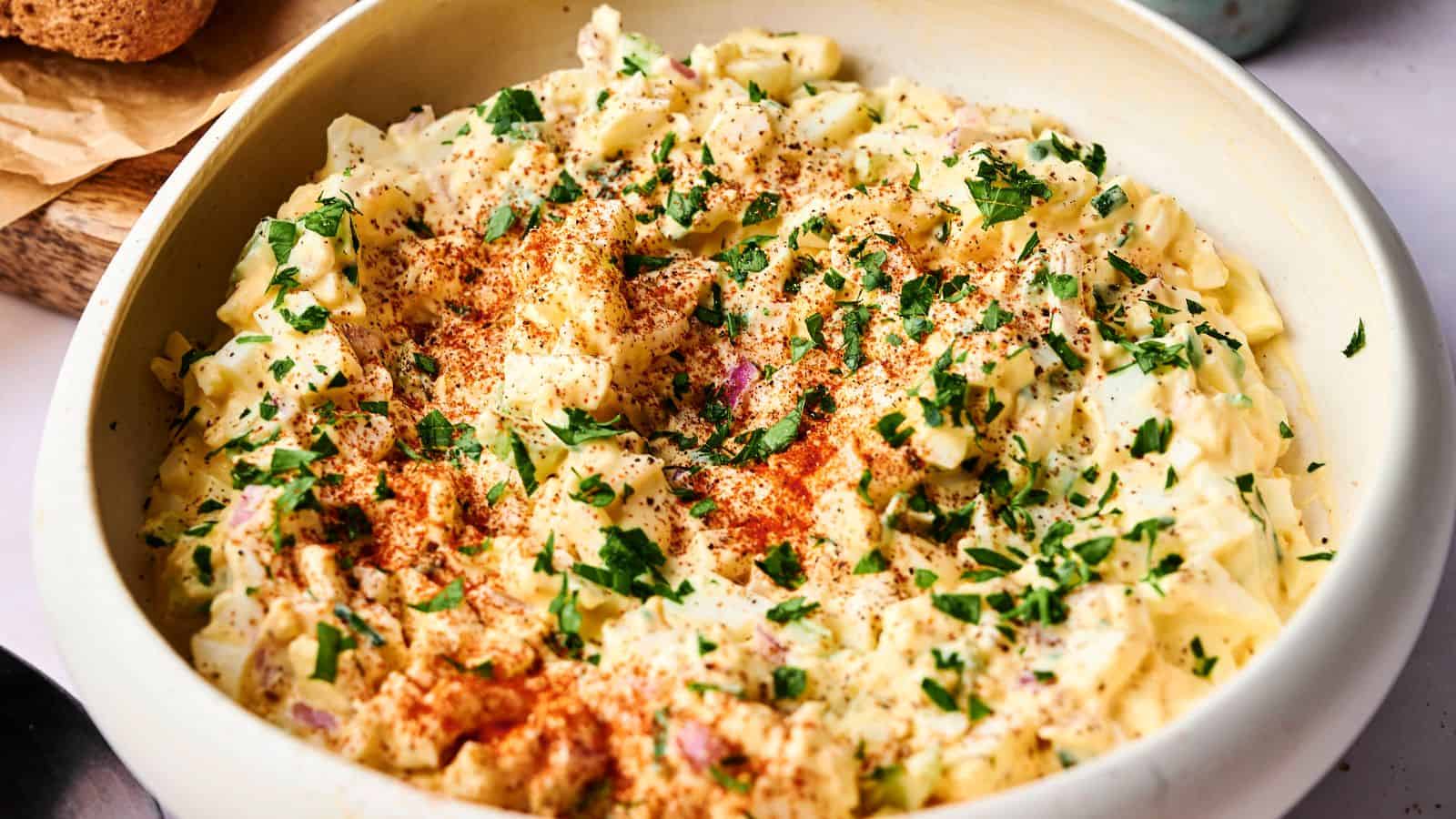 A bowl of creamy egg salad topped with chopped parsley and paprika, set on a light-colored surface.