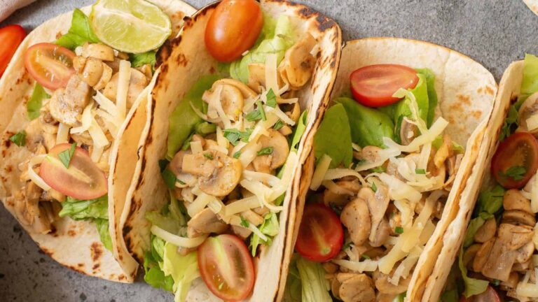 Three soft tacos filled with lettuce, sliced mushrooms, shredded cheese, and halved cherry tomatoes, served with a lime wedge on a gray surface.