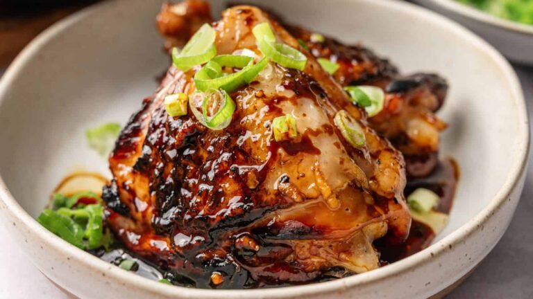 Two pieces of glazed, grilled chicken topped with sliced green onions are served in a white bowl.