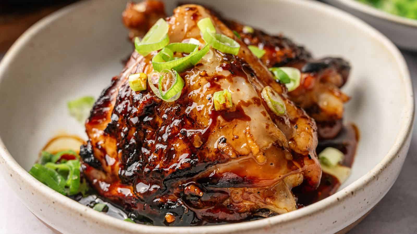 A cooked chicken thigh glazed with a dark sauce, garnished with sliced green onions, served in a light-colored bowl.