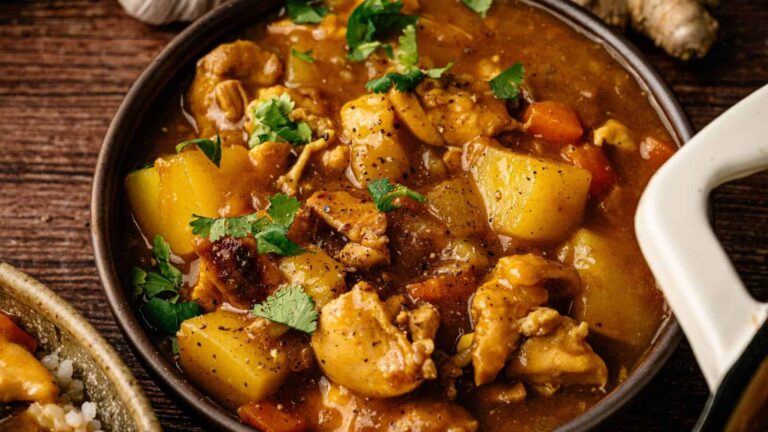 A bowl of stew featuring chunks of chicken, potatoes, carrots, and fresh herbs in a thick brown sauce, placed on a wooden surface.