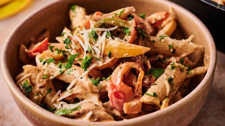 A bowl of penne pasta with creamy sauce, mixed bell peppers, and grated cheese, garnished with chopped parsley.