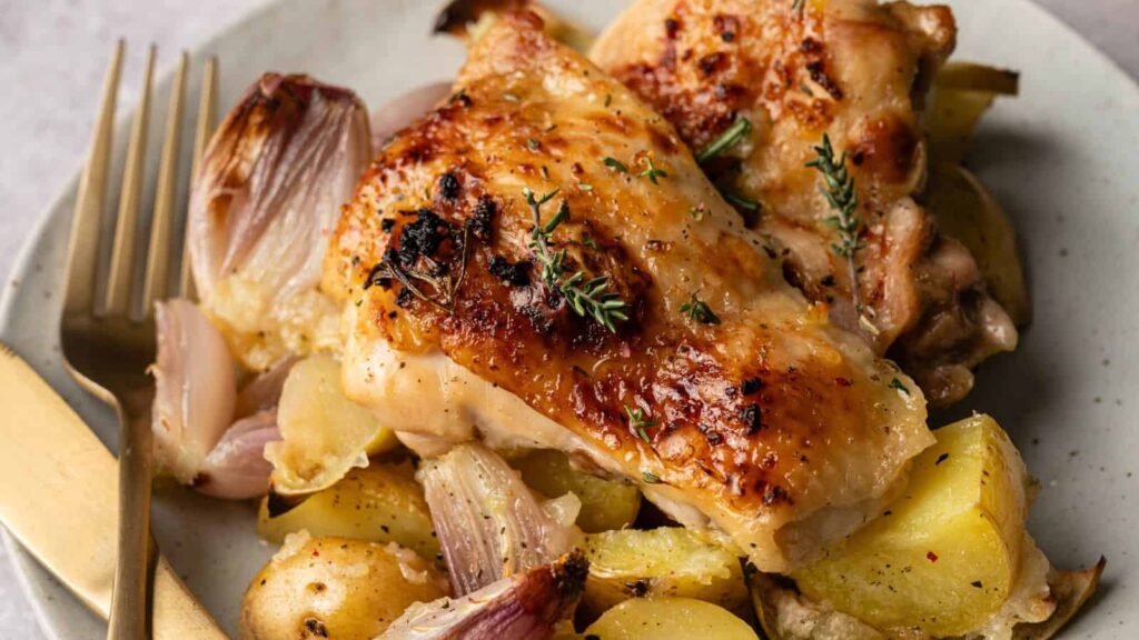 Roasted chicken thighs served on a plate with golden potatoes, roasted onions, and a garnish of fresh thyme, accompanied by a fork and knife.