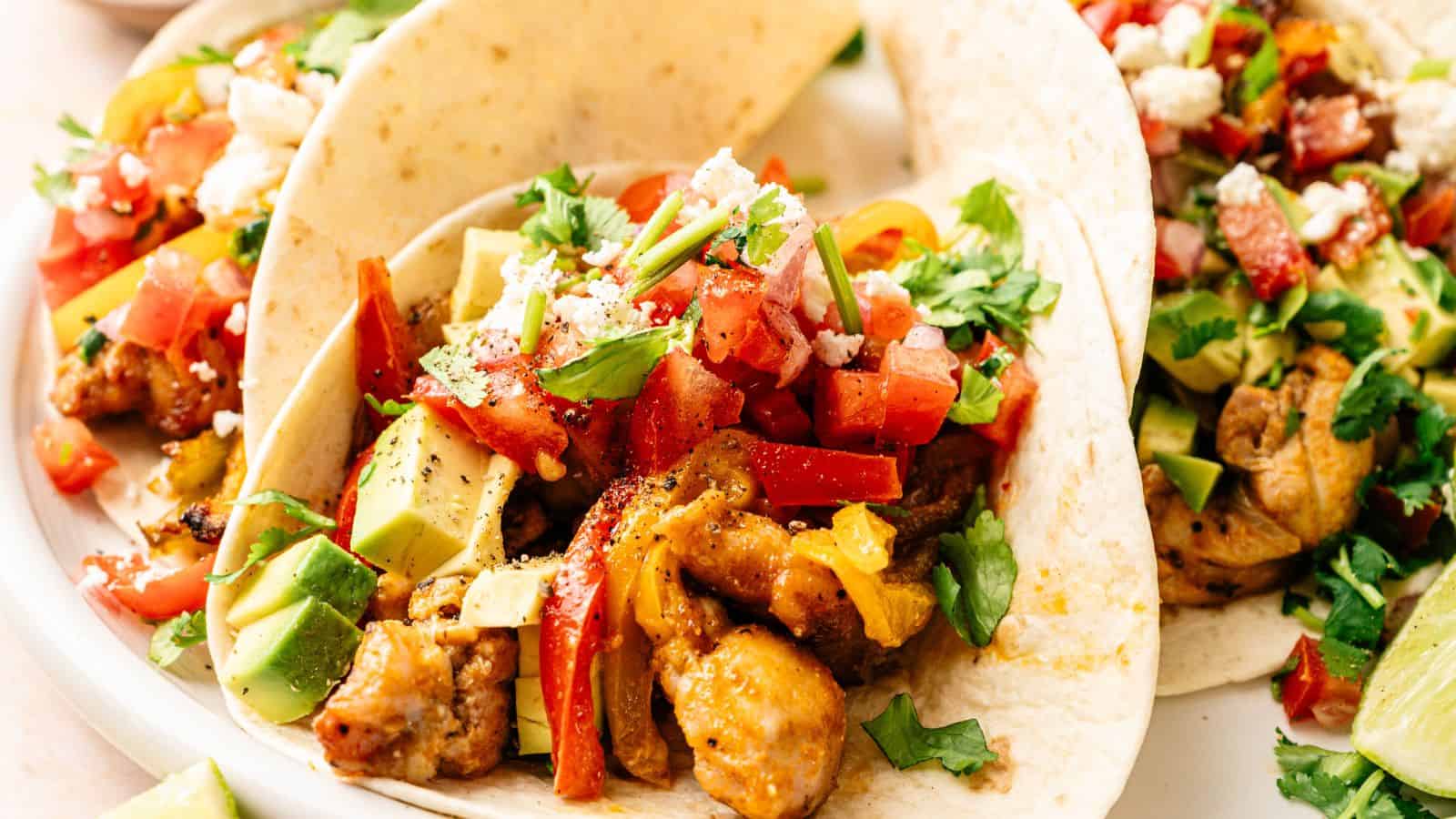 Three soft tacos filled with seasoned chicken, diced avocado, chopped tomatoes, crumbled cheese, and fresh cilantro on a white plate.