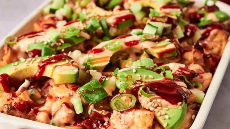 A casserole dish filled with loaded fries topped with avocado slices, diced cucumber, green onions, sesame seeds, and drizzled with creamy sauce and red sauce.