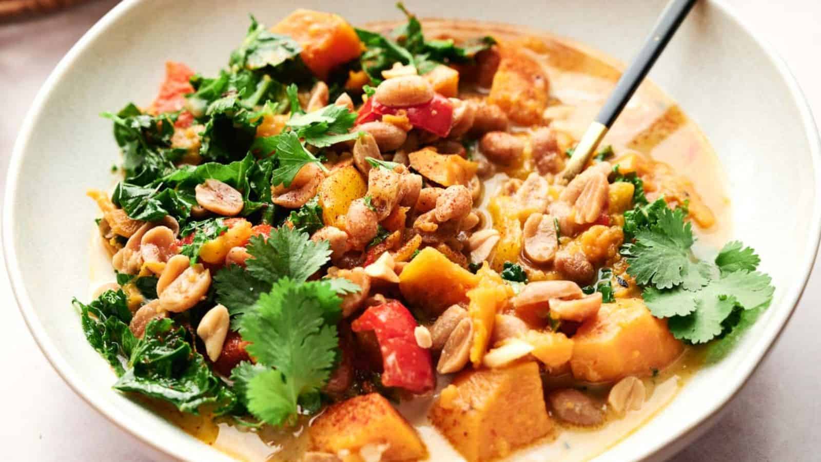 A bowl of vegetable curry with sweet potatoes, kale, red peppers, peanuts, cilantro, and a creamy sauce, served with a spoon.
