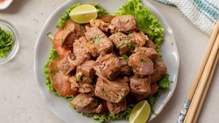 A plate of sautéed beef cubes with onions, served on lettuce and tomato slices, garnished with lime wedges and herbs. Chopsticks are placed to the side.