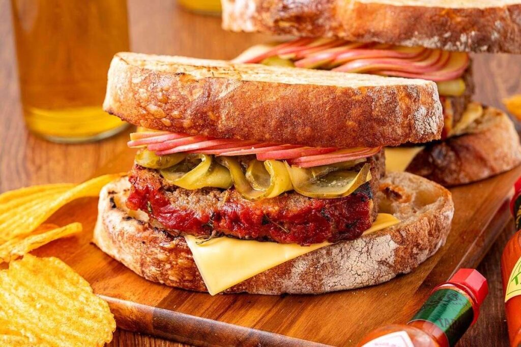 This easy sandwich features sliced bread, cheddar cheese, meatloaf, pickles, and apples, served on a wooden board with potato chips and bottles of hot sauce nearby.