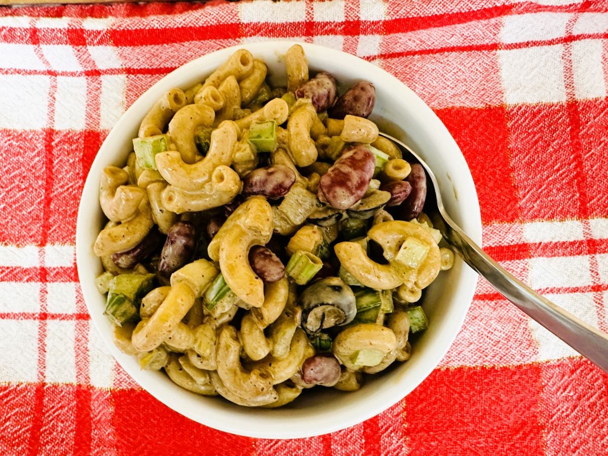 A bowl of Mexican Macaroni Salad with kidney beans, chopped celery, and dressing sits on a red and white plaid tablecloth, with a spoon in the bowl.