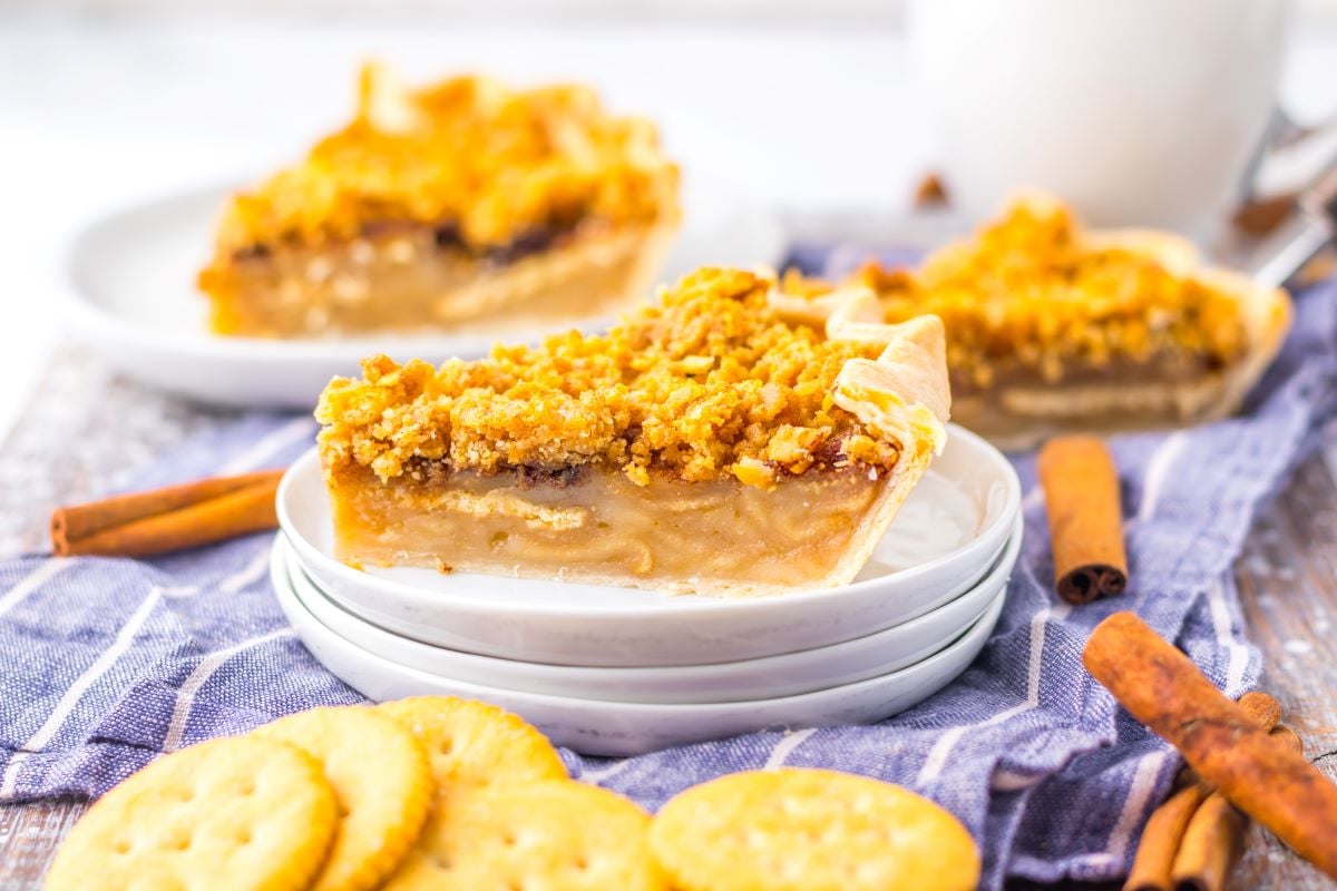 Slice of pie with crumb topping on a stack of plates, surrounded by crackers and cinnamon sticks.