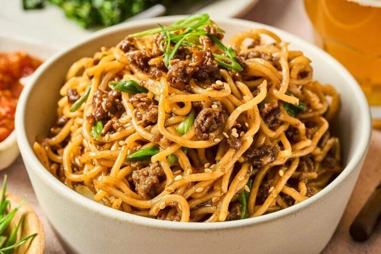 A bowl of noodles mixed with ground meat, sliced green onions, and sesame seeds, served on a table with other dishes in the background—a delicious addition to your collection of flavourful beef recipes.