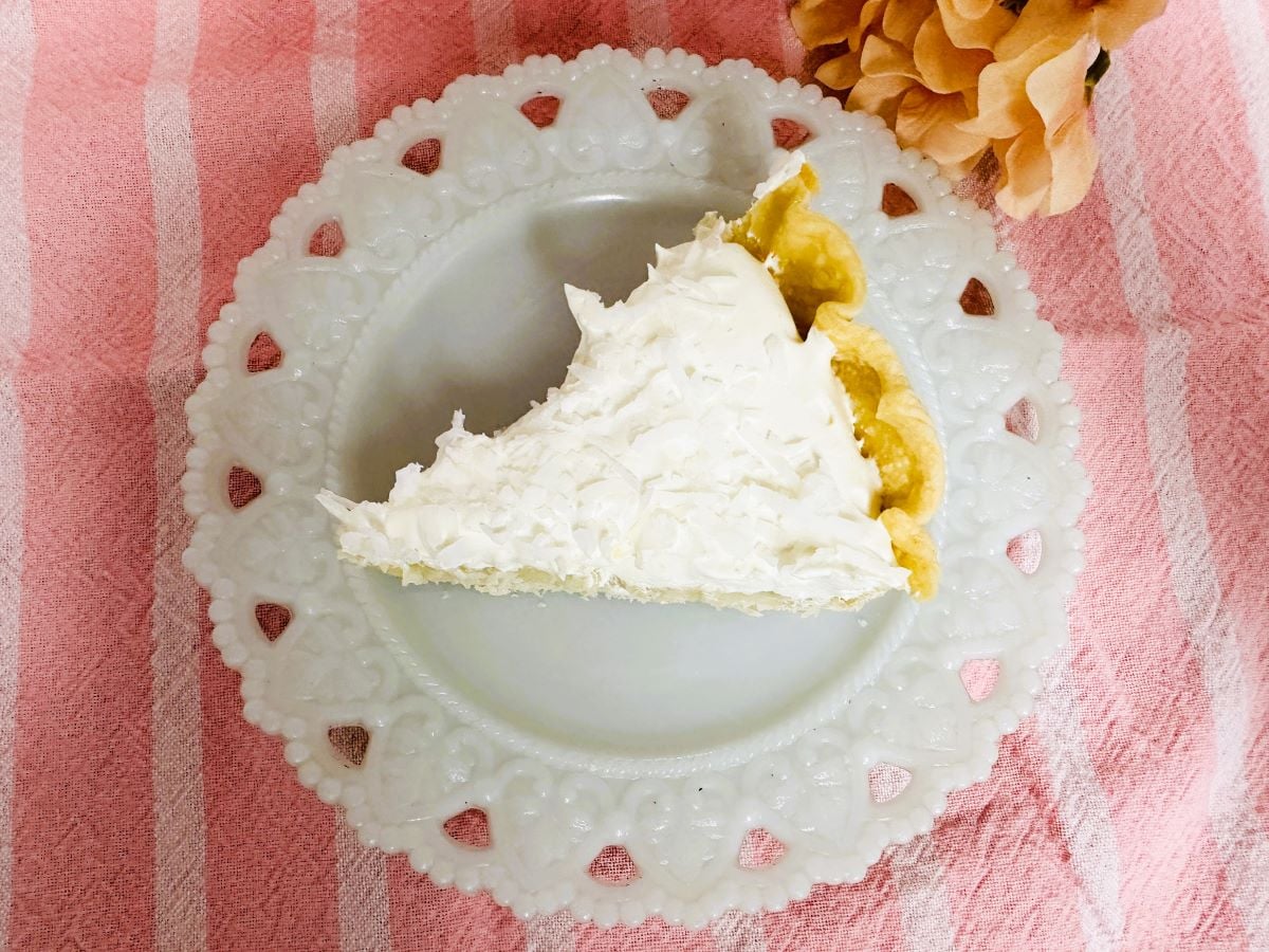 A slice of Old Fashioned Coconut Cream Pie with whipped topping sits on a white decorative plate, placed on a pink striped cloth next to a peach-colored flower.