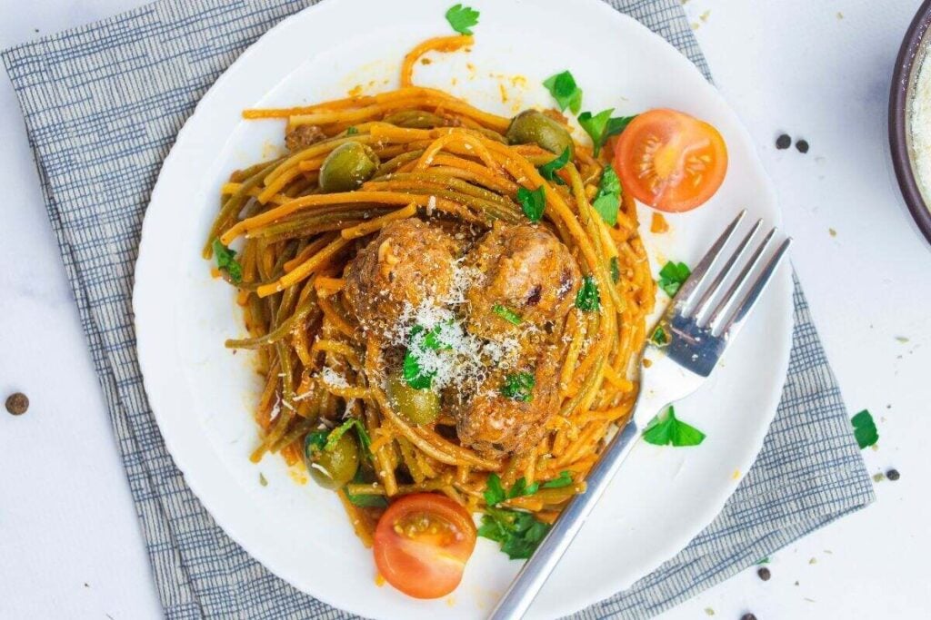A plate of spaghetti with meatballs, green olives, tomato halves, and grated cheese, garnished with parsley, on a white plate with a fork.
