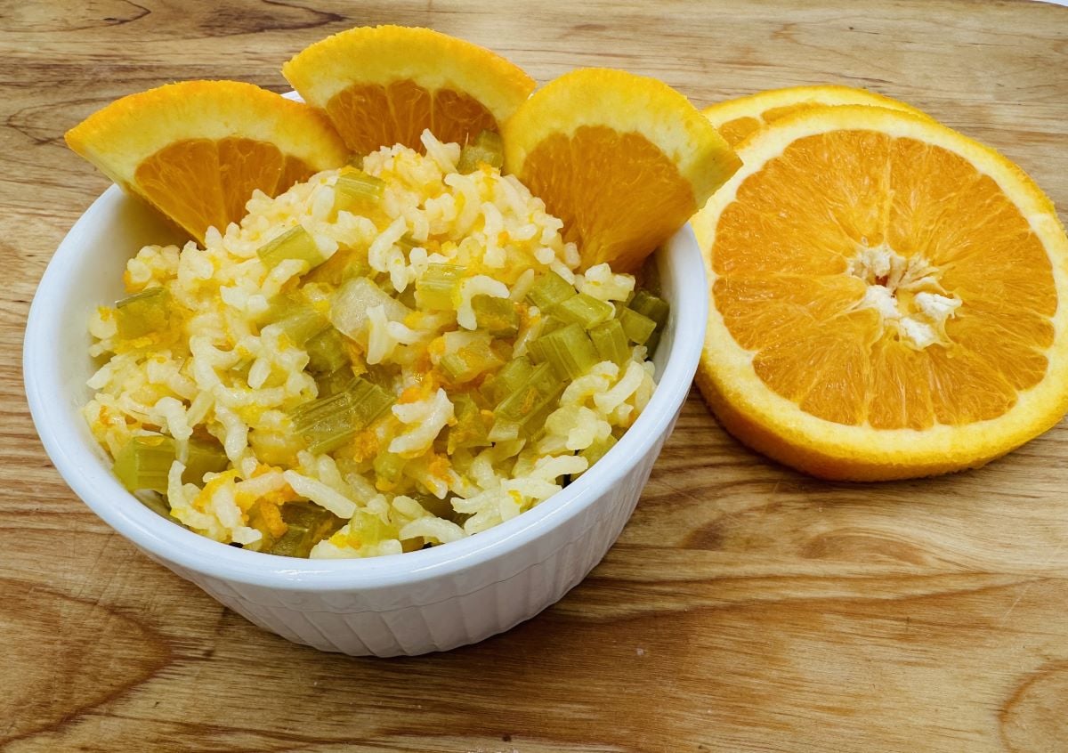 A white bowl filled with rice mixed with chopped celery and orange zest, garnished with orange slices. Two additional orange slices are placed on the wooden surface next to the bowl.