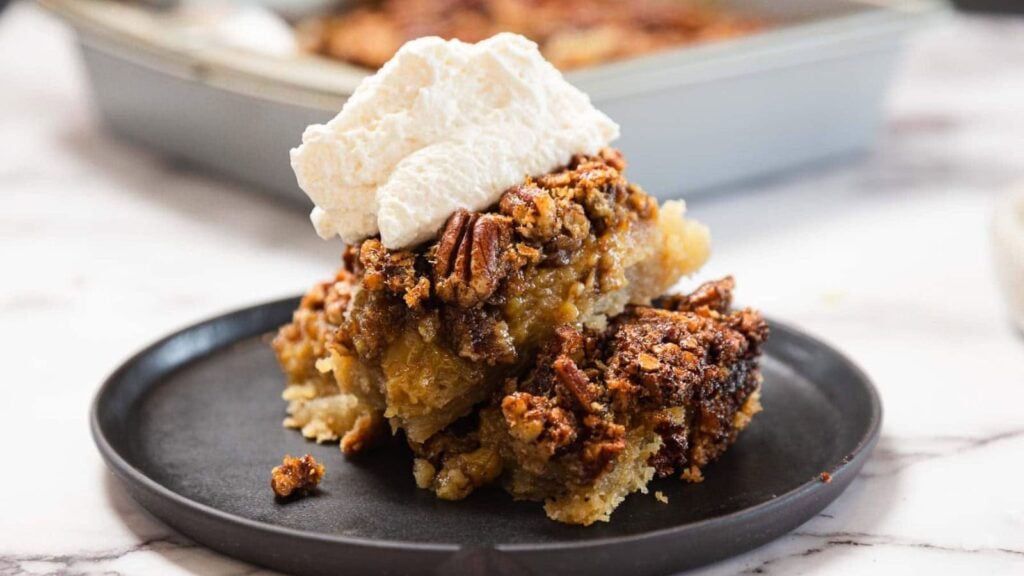 A slice of pecan pie bars topped with whipped cream is served on a black plate, with a baking dish in the background.