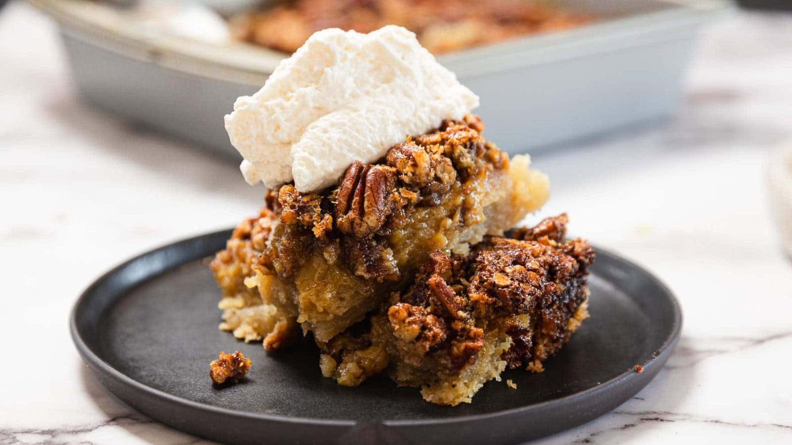 A slice of pecan pie topped with whipped cream on a black plate, with a baking dish in the background.