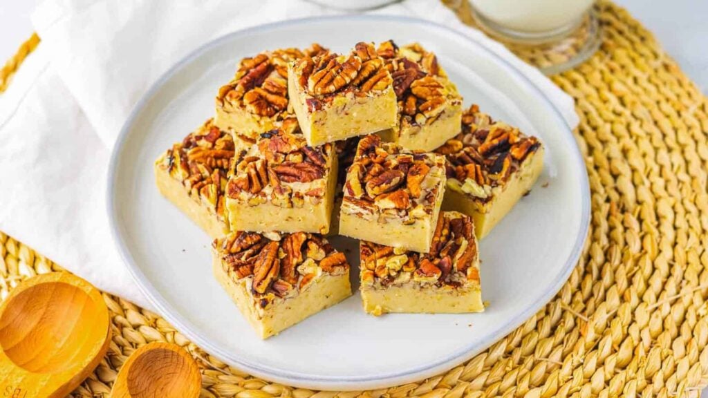 A plate of square pecan-topped dessert bars is arranged on a white plate, placed on a woven placemat with a glass of milk in the background.