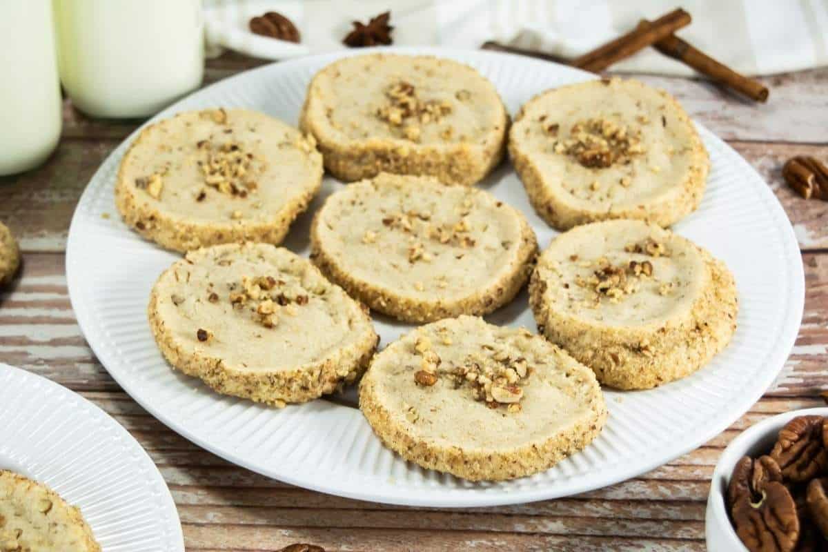 A white plate holds eight round Pecan Shortbread Cookies topped with chopped nuts, placed on a wooden surface next to bottles of milk and cinnamon sticks.