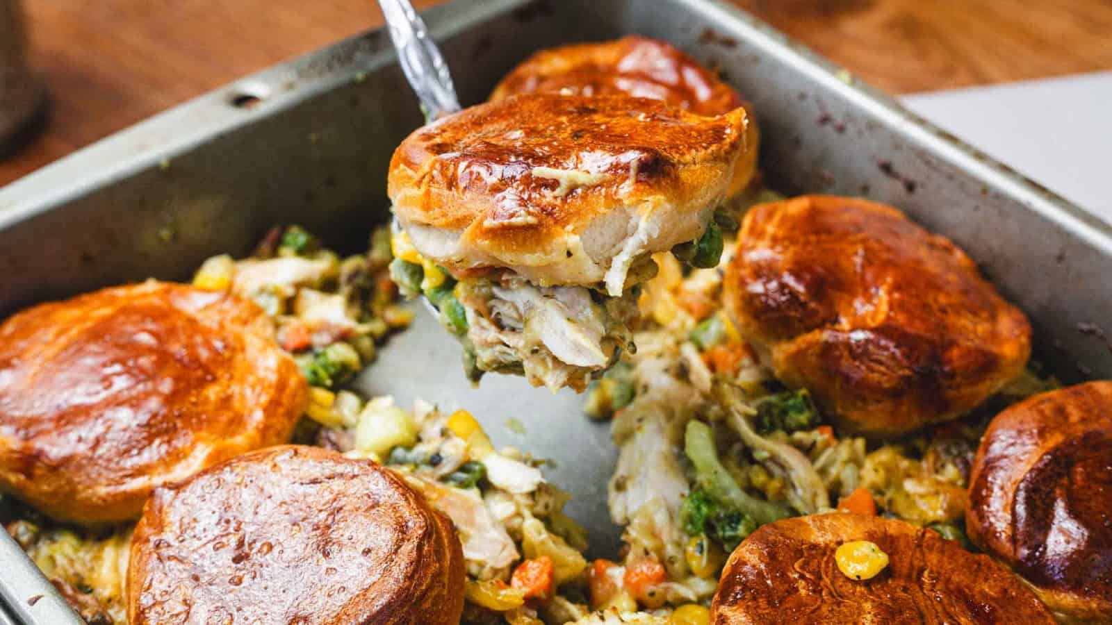 Golden biscuits on creamy chicken pot pie filling with vegetables in a baking pan, one biscuit being served.