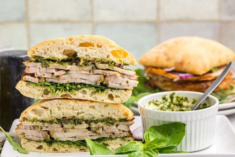 A stacked ciabatta sandwich filled with sliced pork, greens, and pesto is displayed on a plate next to a small white ramekin of pesto and fresh basil leaves—perfect for those seeking easy sandwiches. Another sandwich sits in the background.