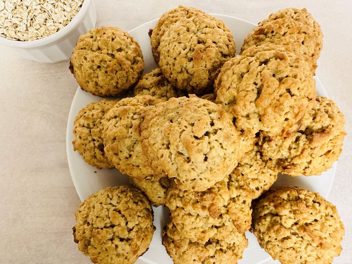 A plate of homemade oatmeal cookies sits on a light surface, with a small bowl of uncooked oats in the upper left corner.