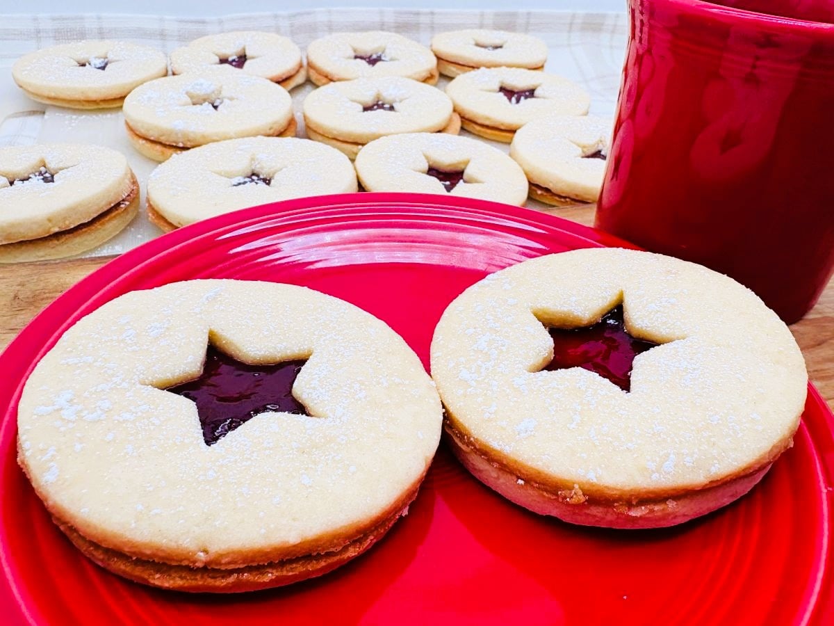 Two round sandwich cookies with star-shaped cutouts revealing jam, on a red plate next to a red mug. More cookies are on a tray in the background.