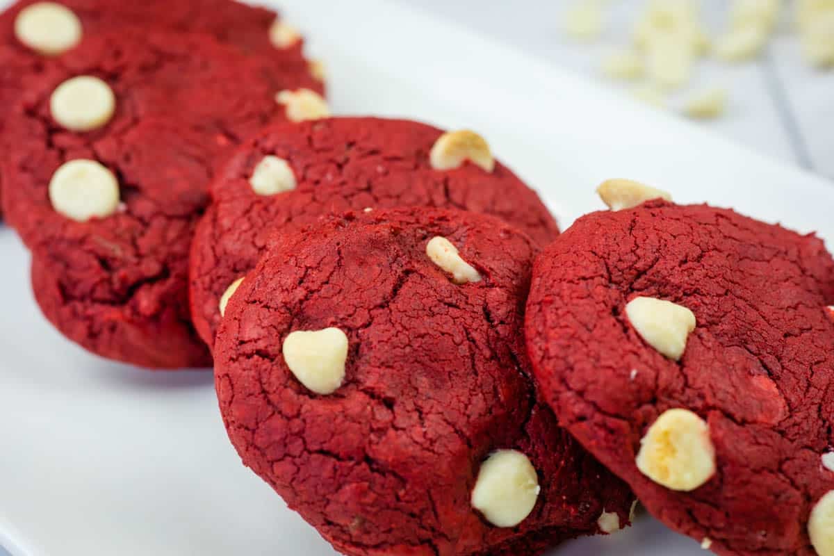 Four red velvet cookies with white chocolate chips on a white rectangular plate.