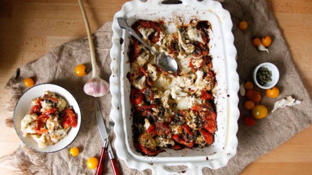 A white baking dish with roasted cauliflower and tomatoes sits on burlap, surrounded by a garlic bulb, cherry tomatoes, capers, and a small plate of the same dish.