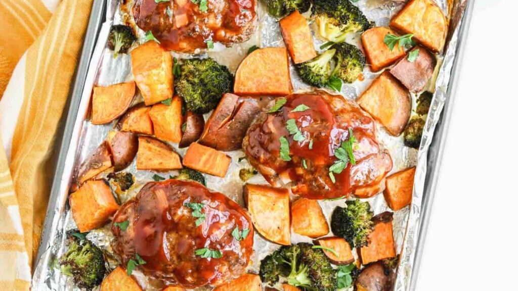 A foil-lined baking sheet with glazed meatloaf patties, roasted sweet potato cubes, and broccoli florets.