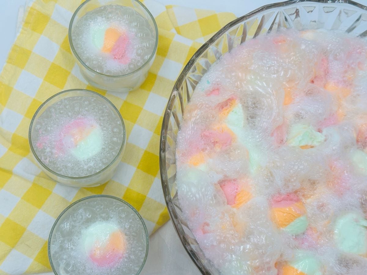 A large glass bowl and three glasses filled with fizzy pastel punch and colorful marshmallows, set on a yellow and white checkered cloth.