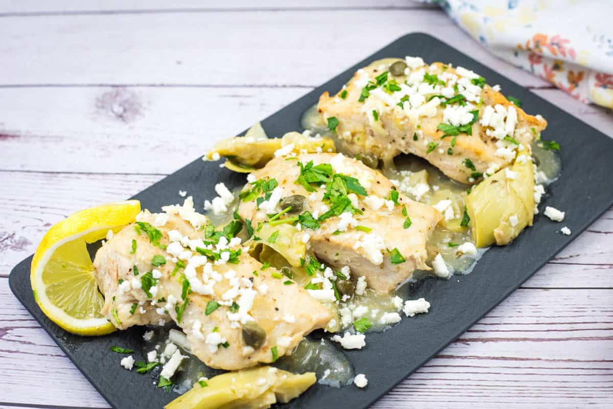 A slate platter with Skillet Chicken with Lemon, Artichokes & Feta topped with herbs.
