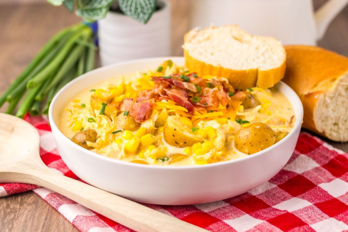 A bowl of creamy potato corn chowder topped with bacon and cheese, served with sliced bread on a red and white checkered cloth.