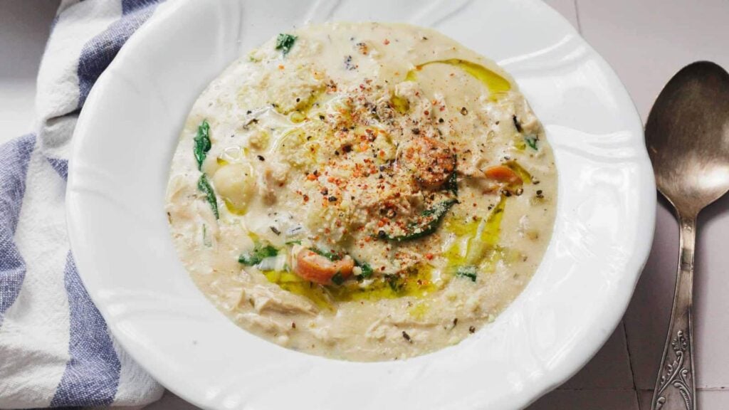 A bowl of creamy soup with vegetables and herbs, drizzled with olive oil and sprinkled with spices, next to a striped towel and a spoon.