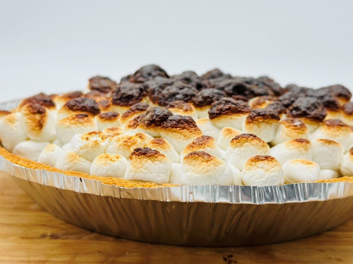 A close-up of a S'mores Pie in a foil pan, topped with toasted marshmallows—some golden brown, others deeply roasted—on a wooden surface with a plain white background.