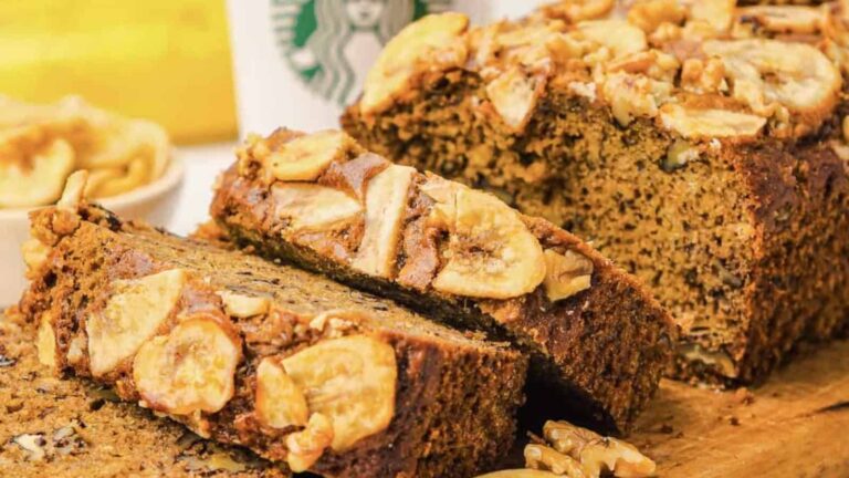 Sliced loaf of banana bread topped with walnuts and banana chips, with a Starbucks cup and a bowl of walnut pieces in the background.