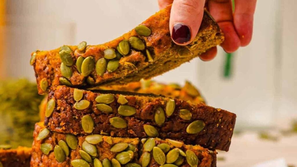 A hand picking up a slice of pumpkin bread topped with pumpkin seeds from a stacked loaf.