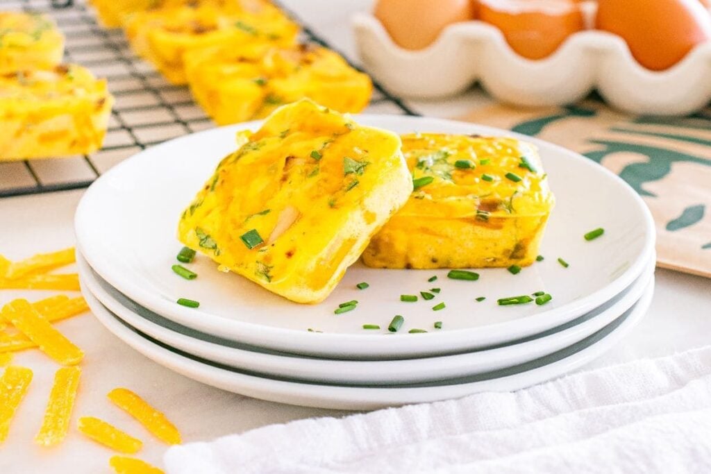 Two square egg bites garnished with chopped chives sit atop stacked white plates, for a perfect copy cat takeout treat. In the background, eggs in a carton and more egg bites cool on a rack.