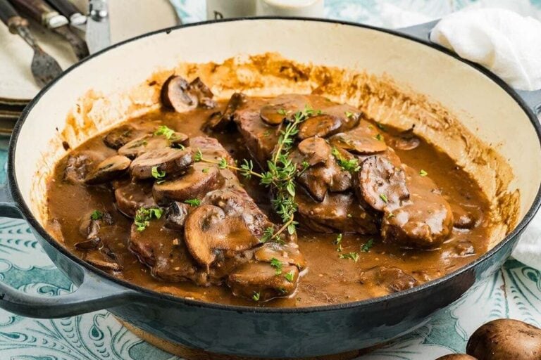 Two pieces of steak covered with brown mushroom sauce and garnished with herbs in a large enameled skillet—perfect for those seeking flavourful beef recipes.
