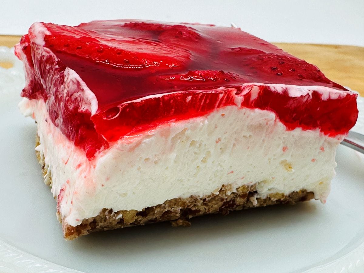 Slice of strawberry pretzel dessert with a layer of strawberries in gelatin, cream cheese filling, and crunchy pretzel crust on a white plate.