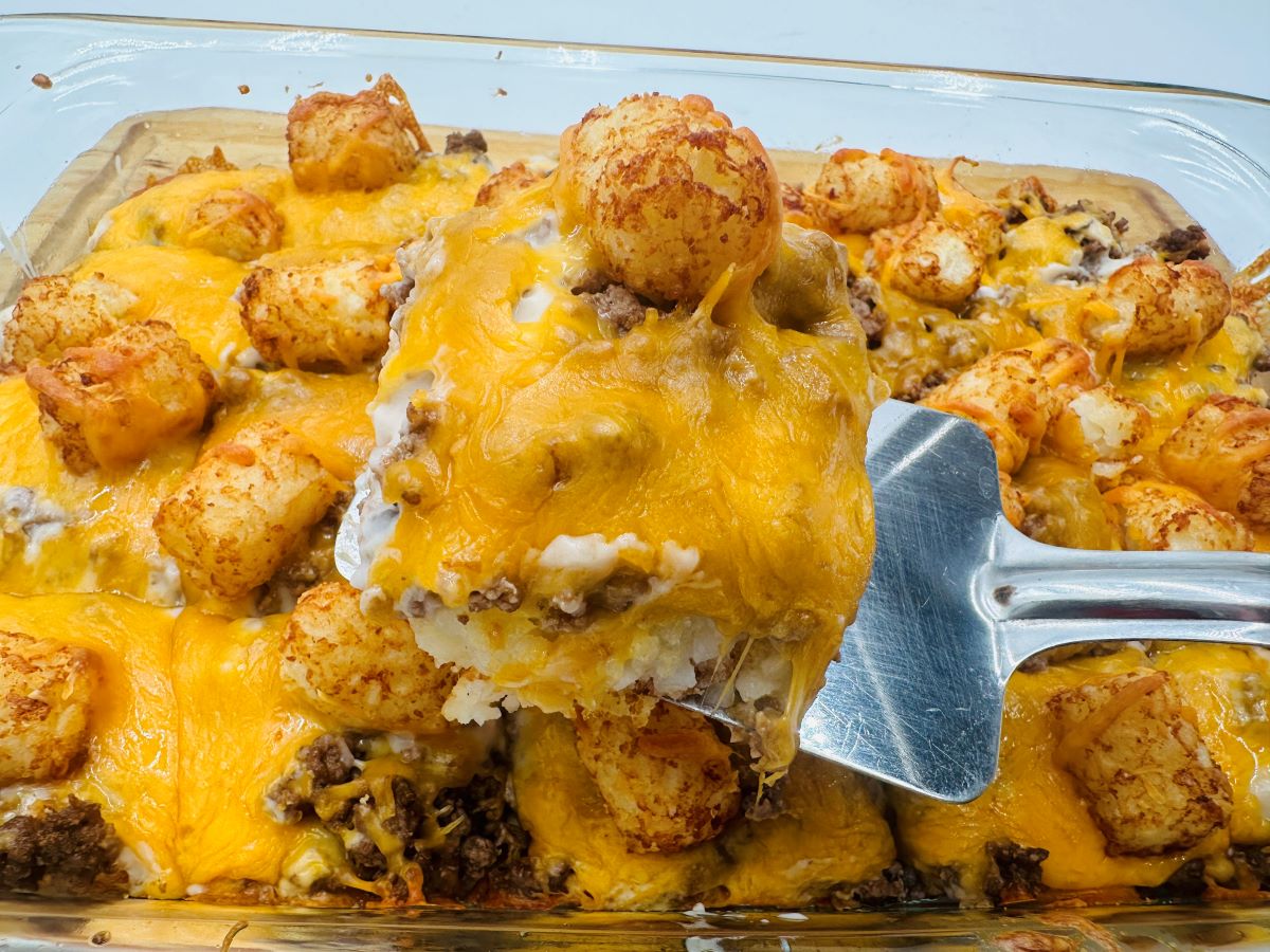 A baking dish of tater tot casserole with ground beef, melted cheddar cheese, and tater tots, with a serving being lifted out on a spatula.