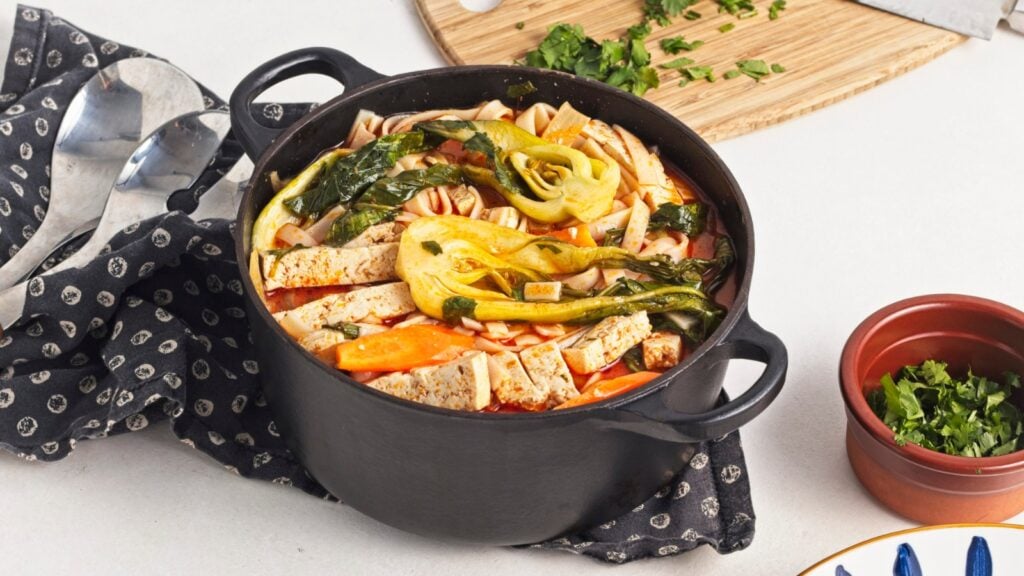 A black pot filled with vegetable soup containing tofu, carrots, greens, and broth, sits on a countertop next to a napkin, spoons, and chopped herbs on a cutting board.