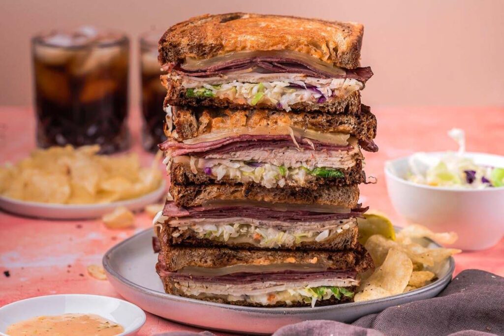 A tall stack of easy sandwiches, classic Reubens layered with meat, cheese, sauerkraut, and sauce on rye bread, served with potato chips and a bowl of coleslaw in the background.