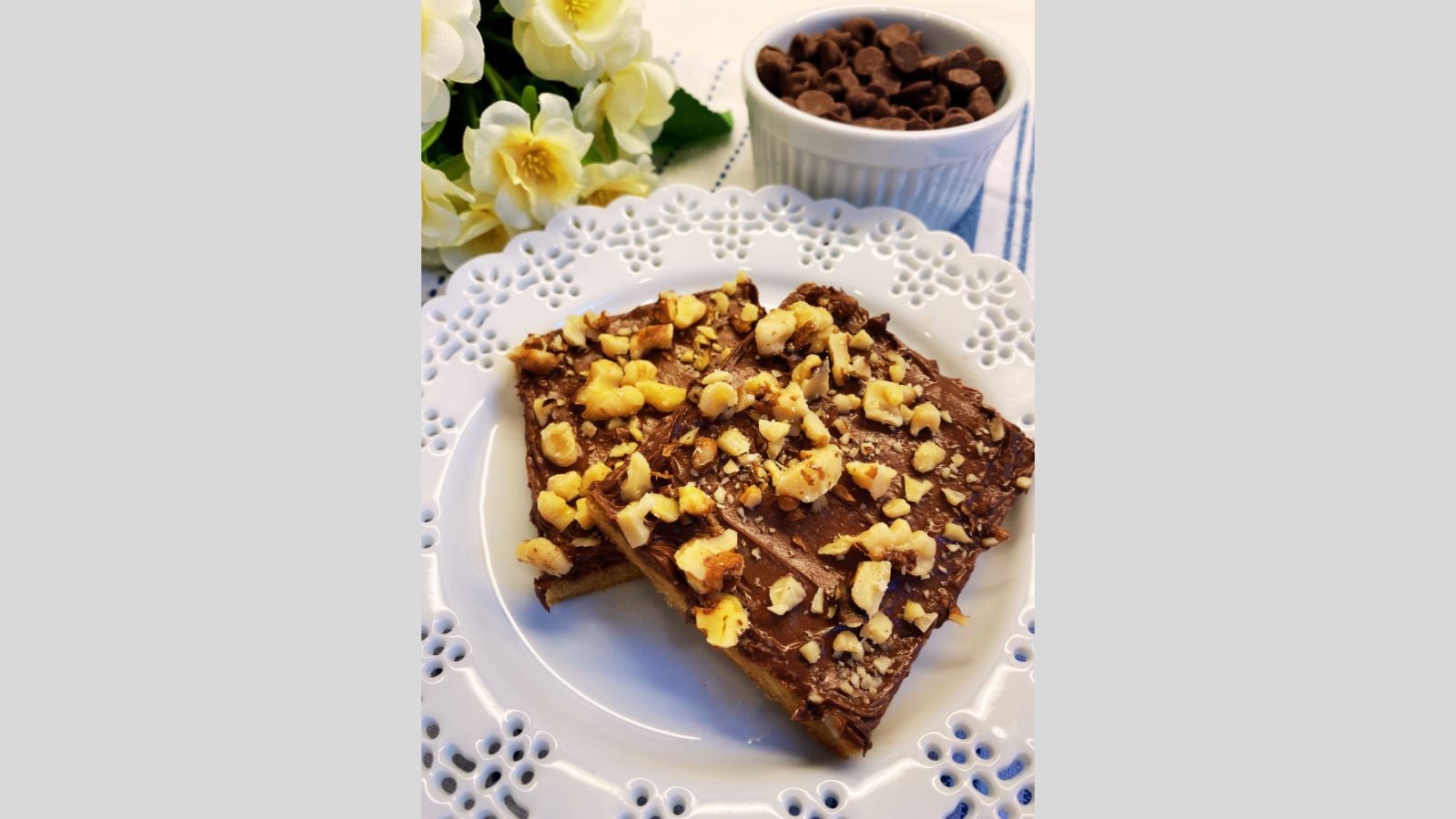 Two pieces of chocolate nut bars topped with chopped nuts on a white plate, with a bowl of chocolate chips and white flowers in the background.