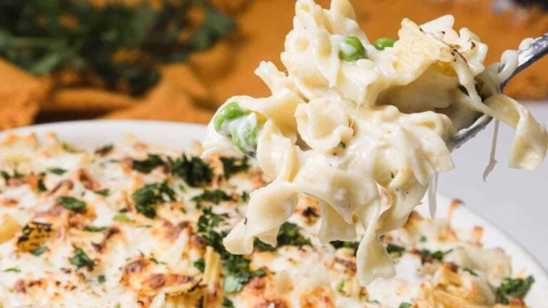 A close-up of a fork holding creamy pasta with green peas above a baked pasta dish topped with melted cheese and herbs.