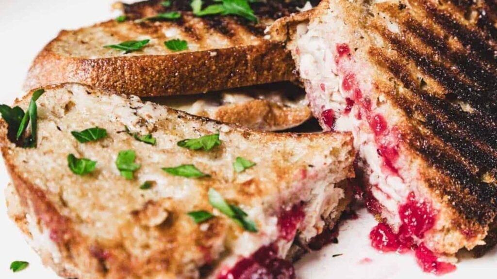 Close-up of a grilled sandwich with toasted bread, visible cranberry filling, shredded turkey, and garnished with chopped parsley.