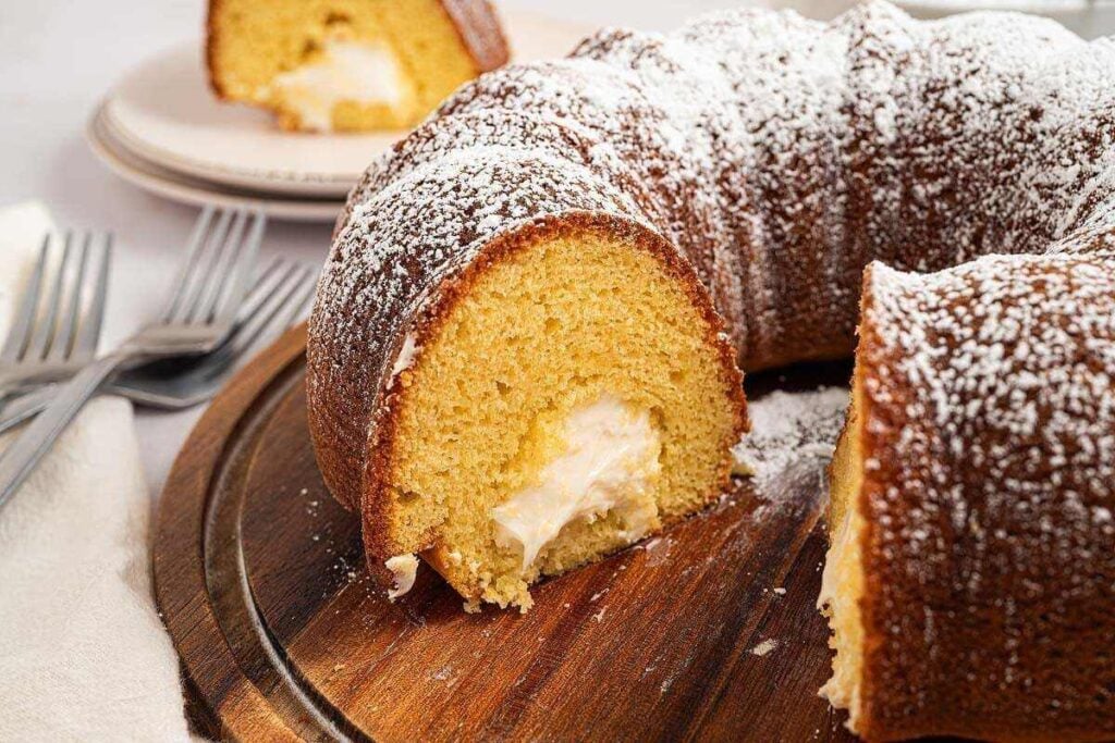 A sliced bundt cake with a cream filling and powdered sugar on top is served on a wooden board, with plates and forks in the background—a perfect treat inspired by tasty cake recipes.