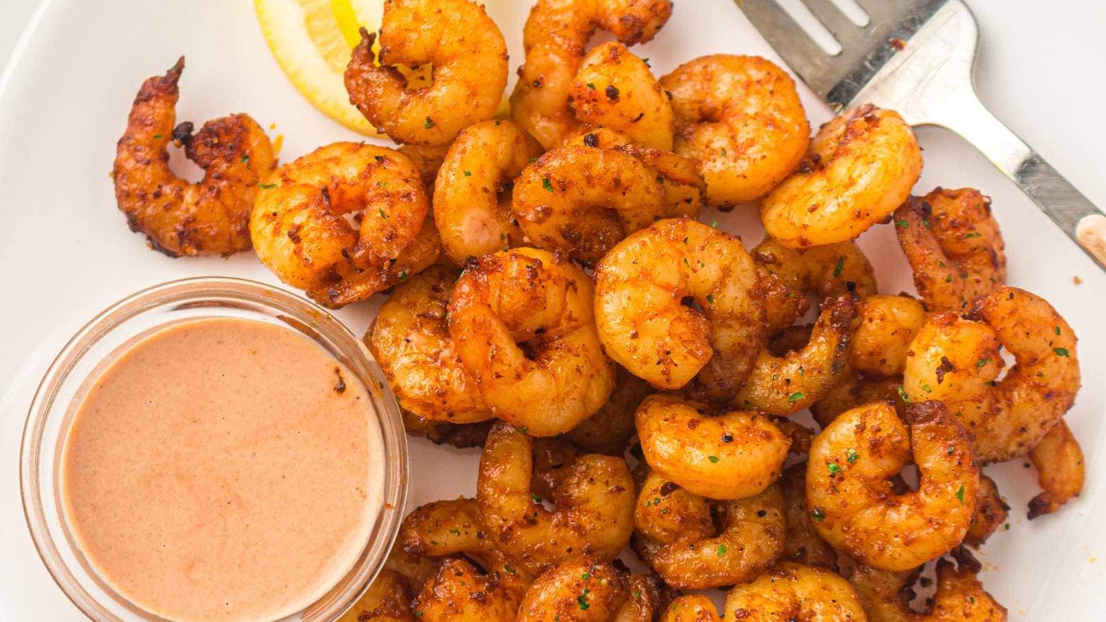 A plate of seasoned, cooked shrimp with a lemon wedge, small jar of dipping sauce, and a metal fork on the side.